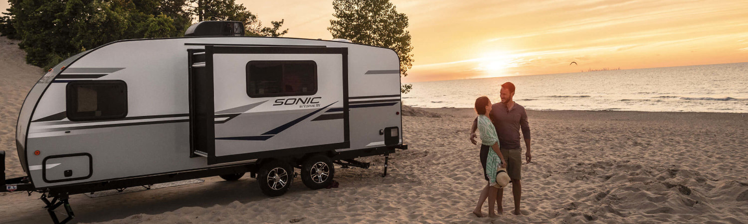 2020 Venture RV Sonic Travel Trailer parked in the sand while a couple enjoy a romantic sunset …